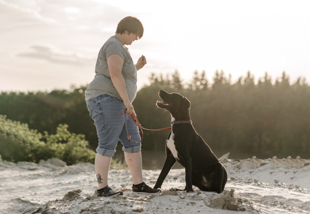 Frau steht gegenüber eines Hundes, beide sehen sich an, sie befinden sich am Strand